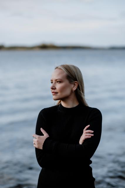 Porträtt på Matilda Svenfelt med havet i bakgrunden. Hon är iklädd en svart, långärmad, figurnära klänning och håller armarna i kors. Tittar mot vänster, så man ser hennes profil. Hon har utsläppt rakt, blont hår.